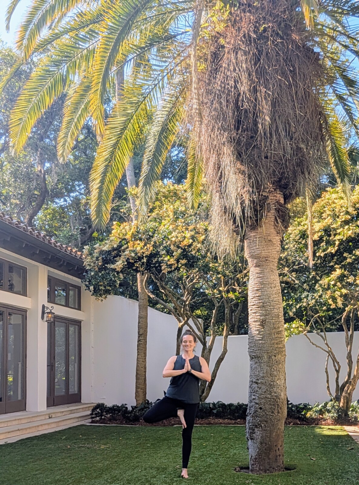 Woman with brown hair dressed in athletic clothing stands in tree pose next to a tree.