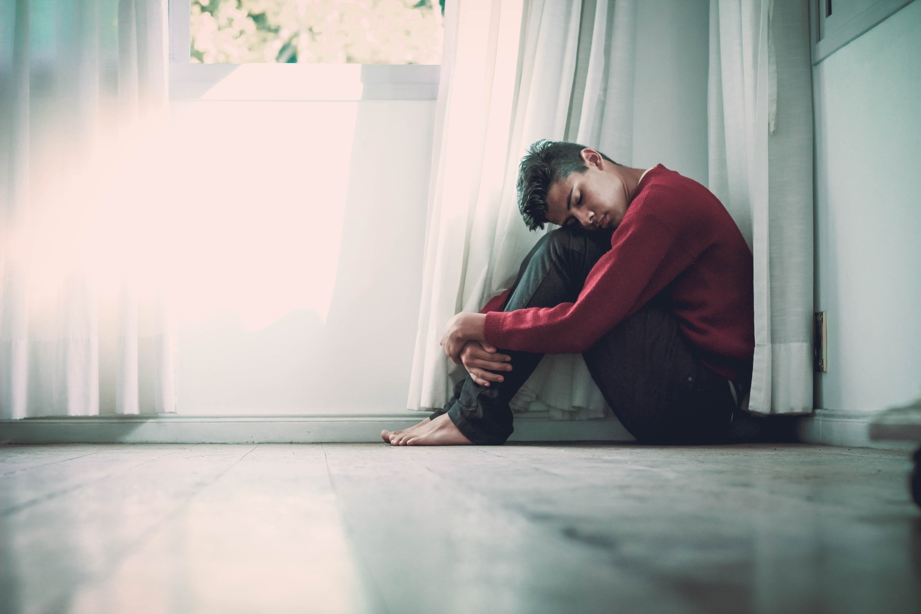 An Indian man sitting on the floor, his knees are pulled to his chest, his head is resting on his knees and his eyes are shut.