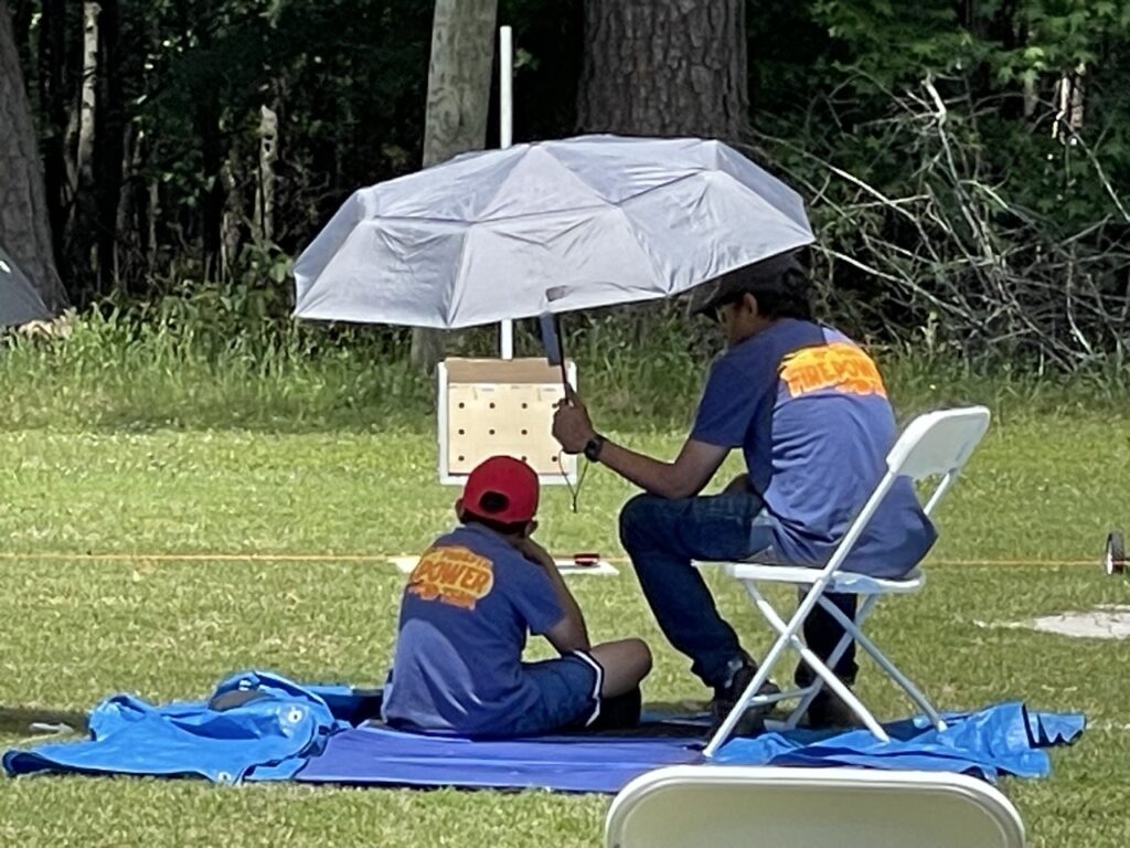 A parent holds an umbrella over his 6th grade son while the youth performs a sitting BB shot.