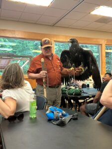 An adult man has an adult golden eagle sitting on his arm as he presents information to a classroom of people.