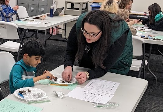 An adult leans over a table where an elementary school student investigates an owl pellet.