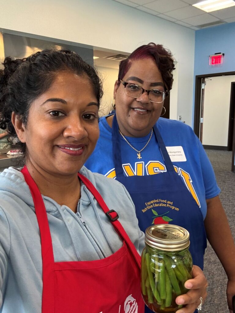 Two adult women, wearing aprons, one is holding a jar of pickled green beans