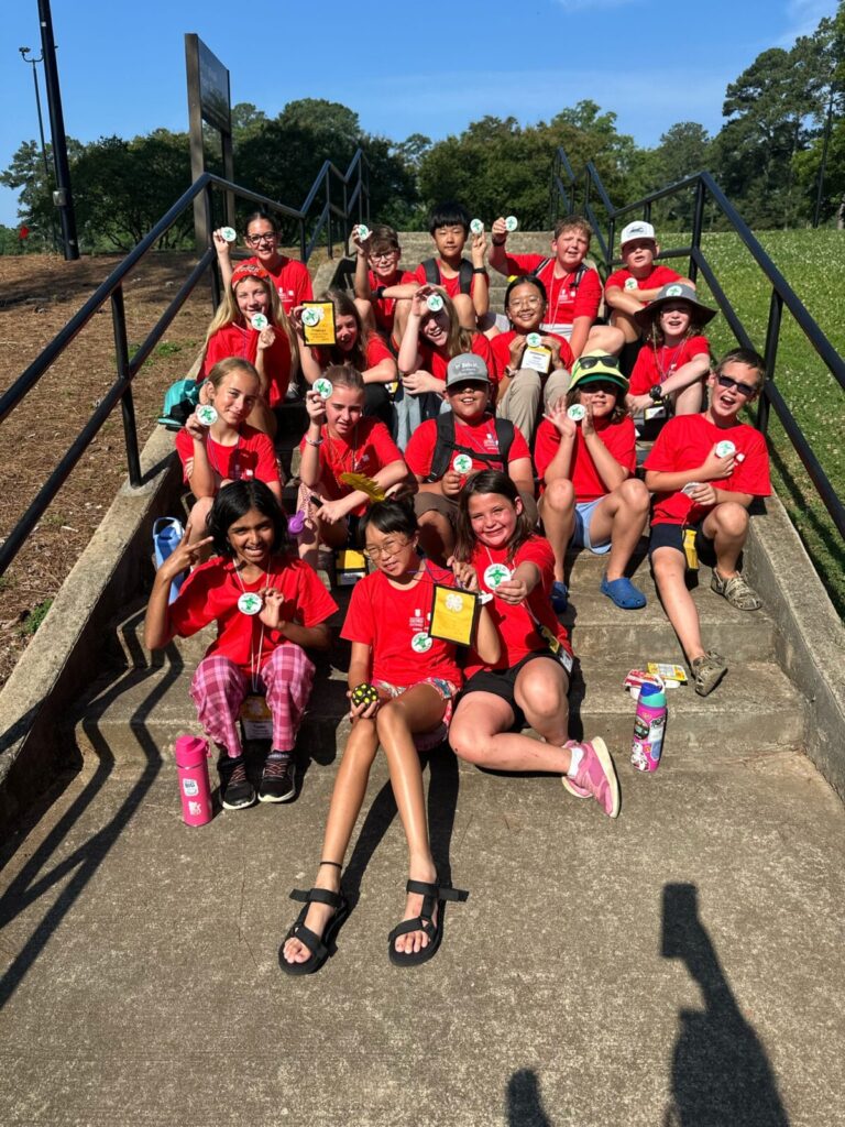 Students dressed in matching red camp shirts sitting on the stairs showing off their winning camp buttons.