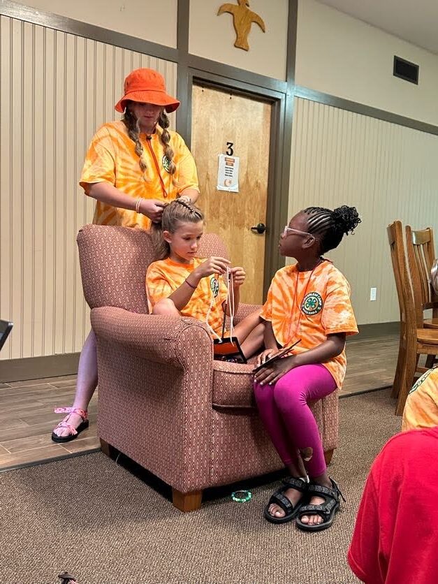Two students sitting in a chair. One is getting her hair braided by a teen leader and the other is watching.