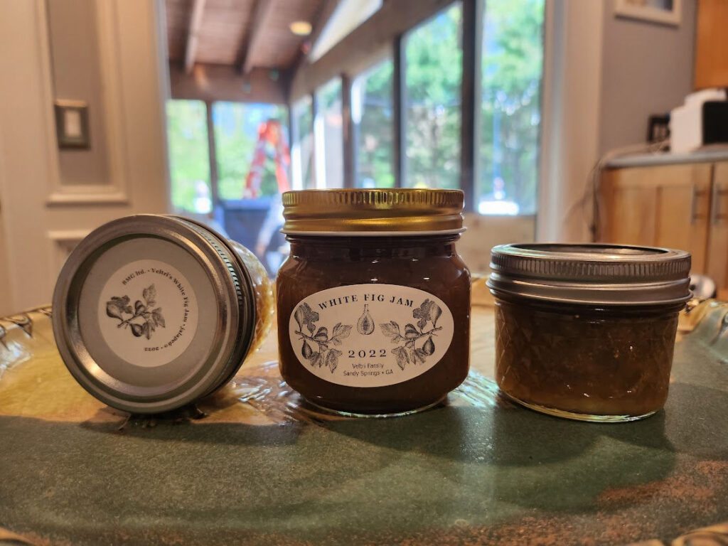 Three glass jars of homemade white fig jam with decorative labels sit on a decorative green tray, against a cozy, sunlit kitchen backdrop.