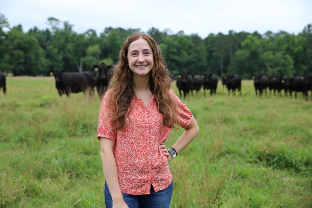 A woman stands in a grassy field with a herd of black cows in the background.