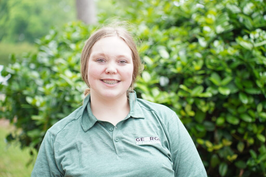 teenage girl wearing a green Georgia 4-H Ambassador polo standing next to a tree