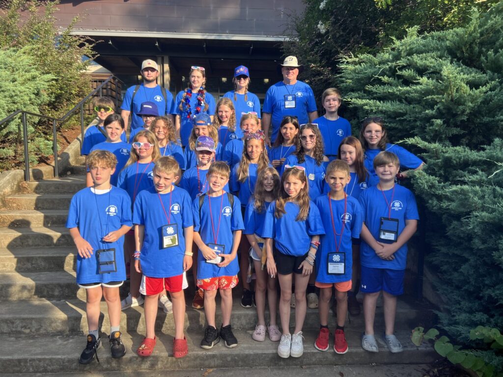 Twenty-seven campers wearing blue camp shirts posing on steps surrounded by tall green bushes.