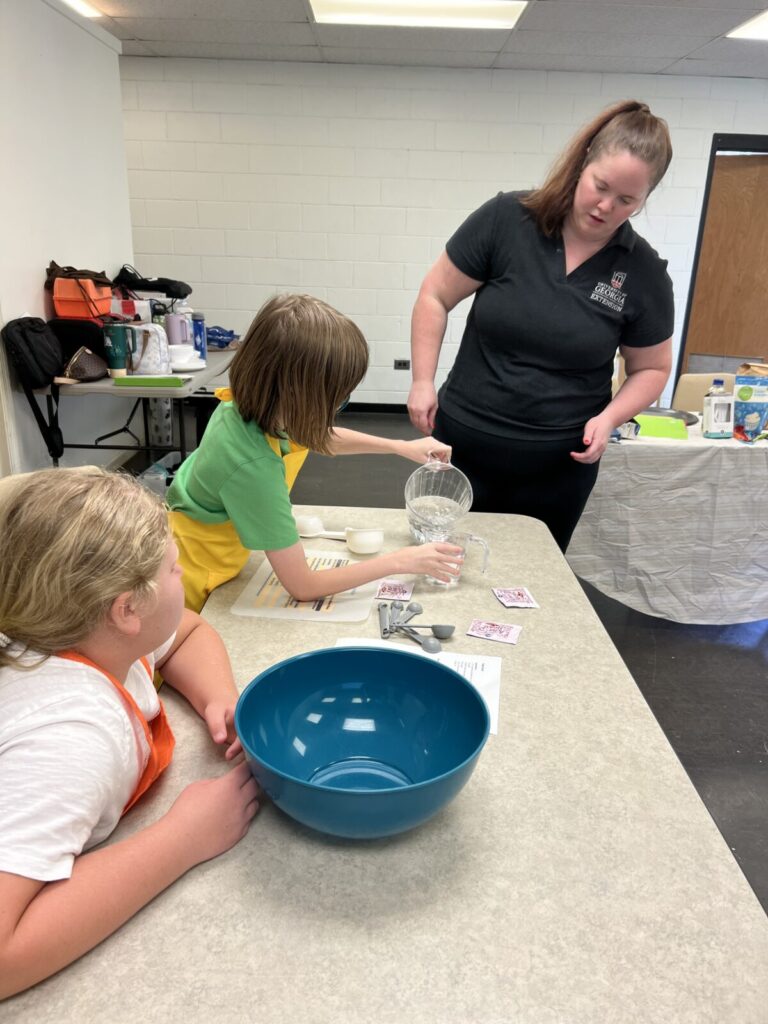 Adult woman standing near two young girls as they measure ingredients for a recipe