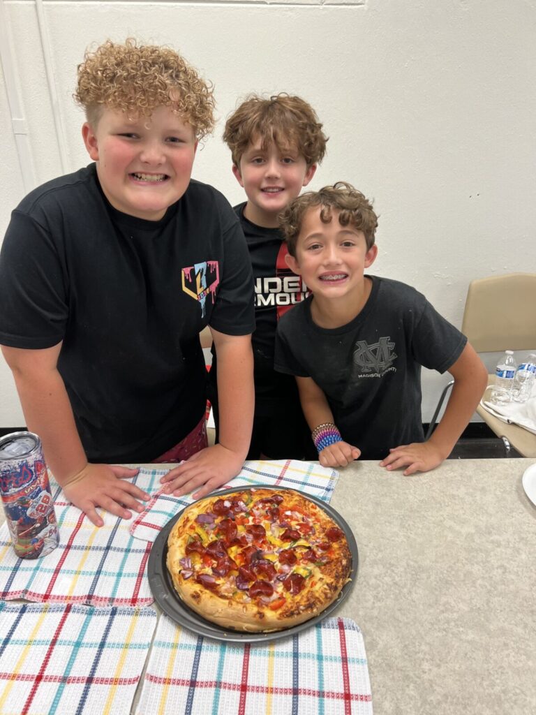three young boys smiling while standing in front of pizza