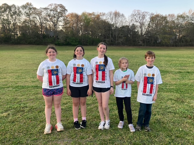 Five youth wearing matching t-shirts with state of Georgia flag as they stand in a field of grass