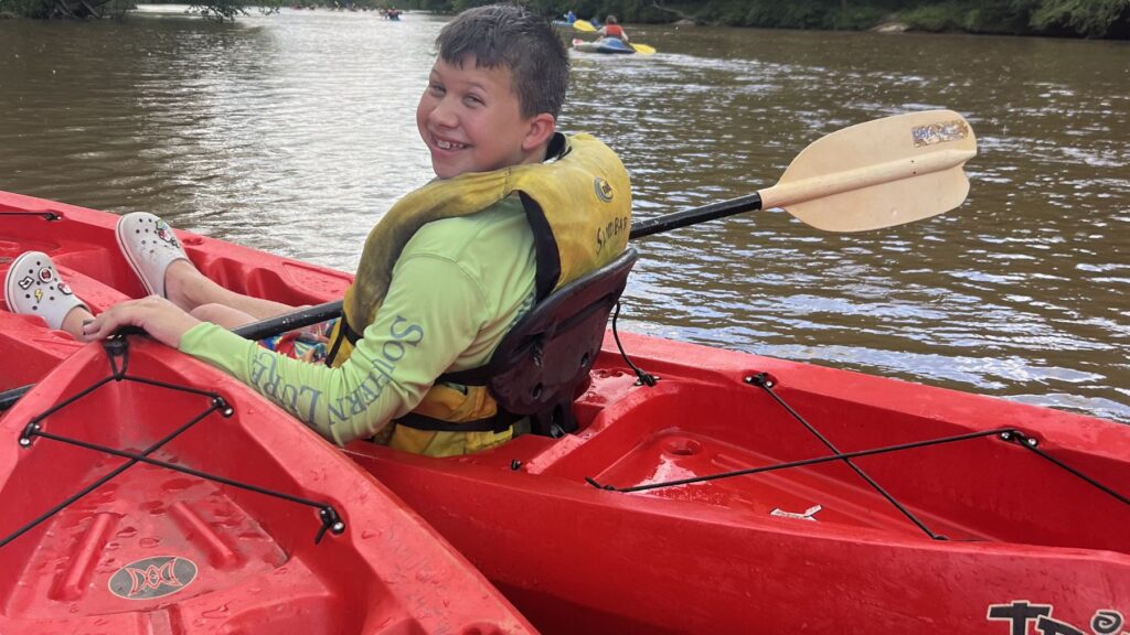 A person in a yellow life vest and lime green shirt smiles while sitting in a red kayak on a river, holding a paddle.