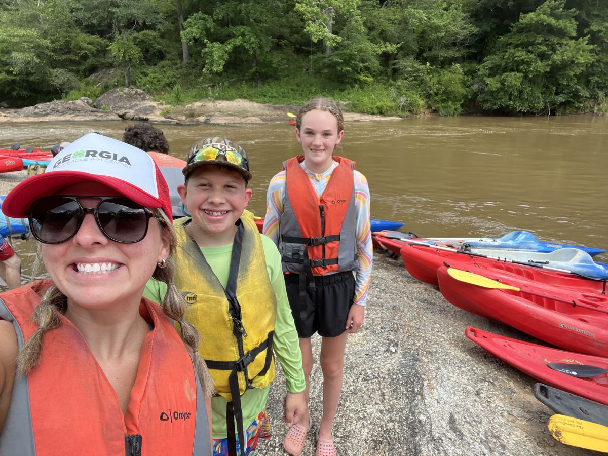 A group of people in life vests stand by a river with kayaks lined up on the shore.
