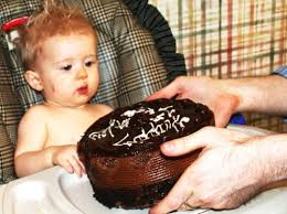 Toddler in high chair looking at a chocolate birthday cake held out to them.